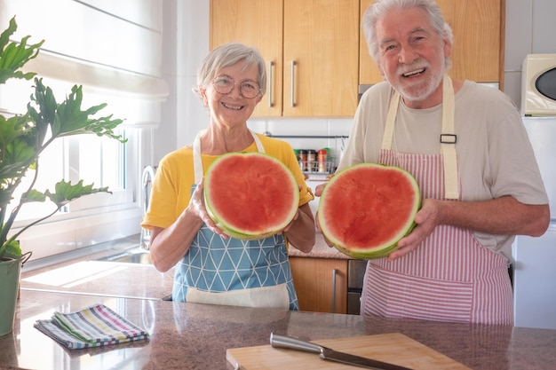 Happy senior caucasian couple in home kitchen holding a big and heavy watermelon cutted in a half looking at camera smiling hydratation freshness diet and healthy eating concept