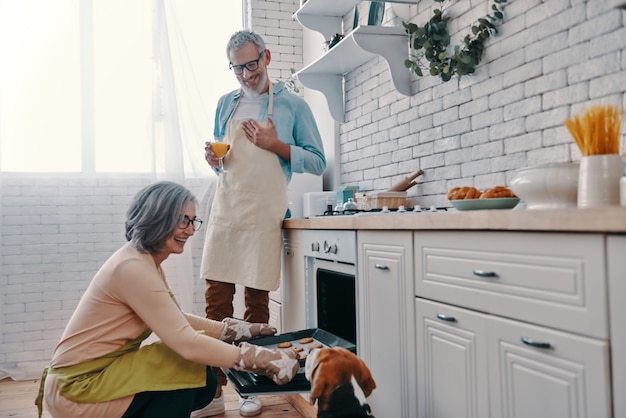 Happy senior couple in aprons preparing healthy dinner in oven and smiling while spending time at home