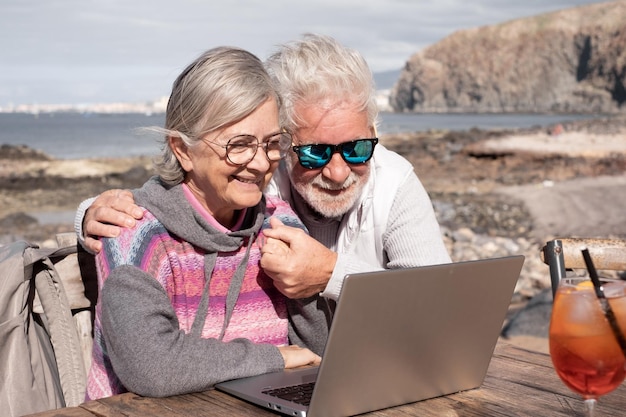 Happy senior couple browsing together on laptop enjoying sunny day outdoor at the beach