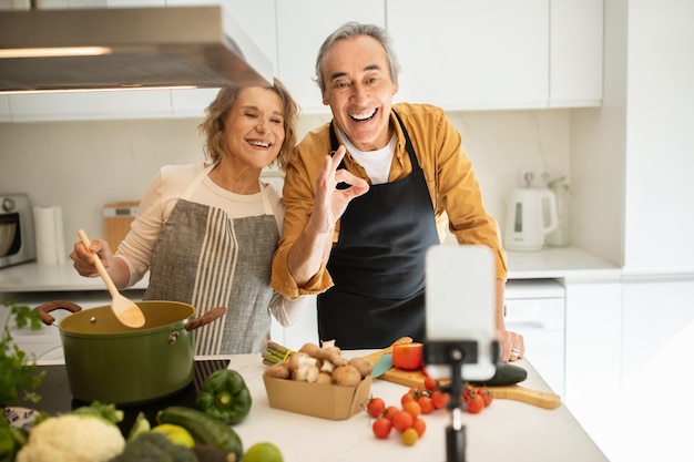 Happy senior couple food bloggers streaming while preparing dinner at home showing ok gesture modern