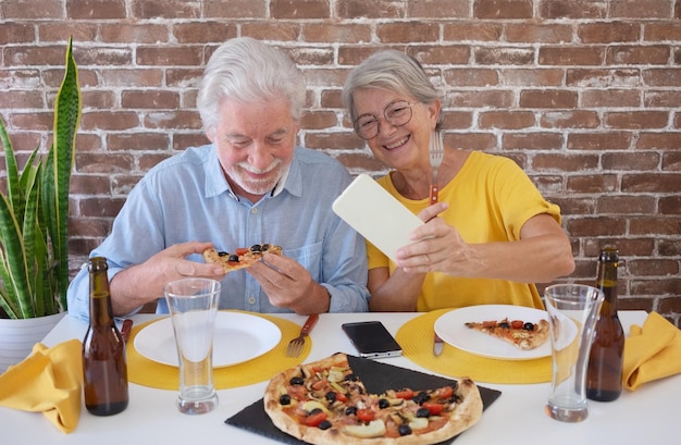 Happy senior couple having fun together with pizza and beer Elderly woman and man laughing sitting at home table using phone for a selfie