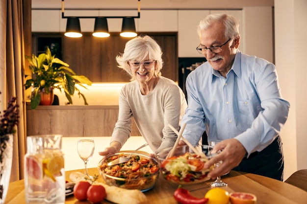 A happy senior couple is putting food on lunch table at home