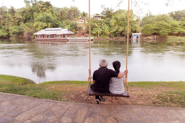 Happy senior couple sitting on swing together near river waiting for sunset.