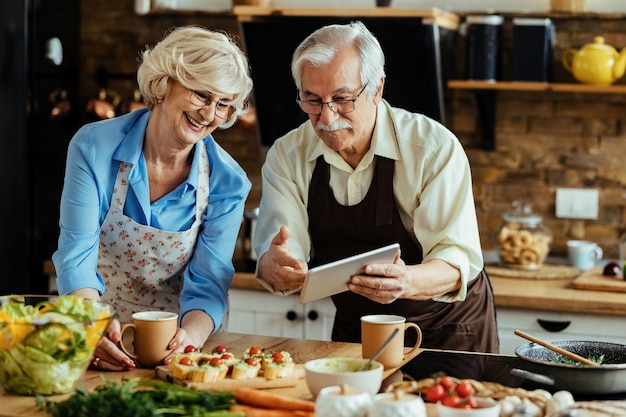 Happy senior couple surfing the net on touchpad while preparing food in the kitchen