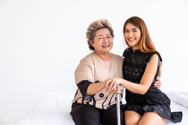 Happy senior elderly Asian woman female mother holding cane with daughter sitting on bed in bedroom looking at camera