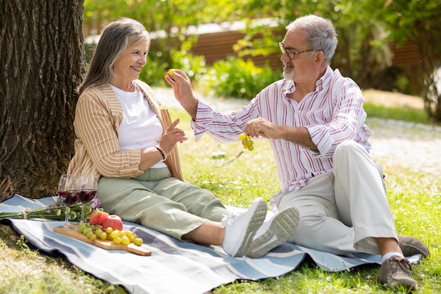 Happy senior european man feeding grapes to lady enjoy romantic date lunch in park have fun