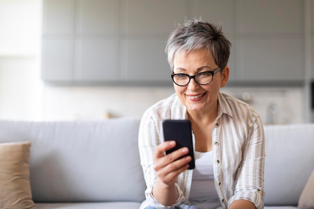 Happy senior lady using mobile phone while relaxing on couch at home