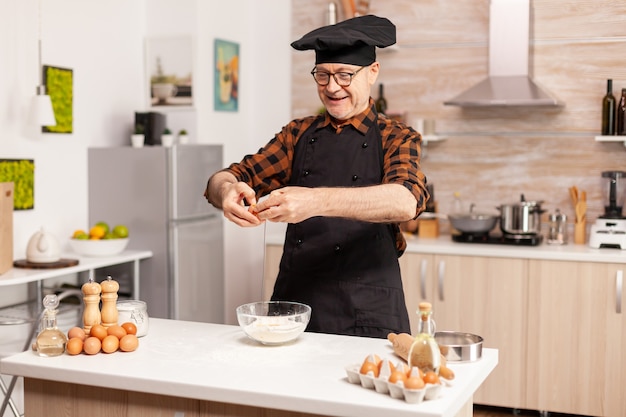 Happy senior man cracking eggs over wheat flour while preparing traditional recipe. Elderly pastry chef cracking egg on glass bowl for cake recipe in kitchen, mixing by hand, kneading