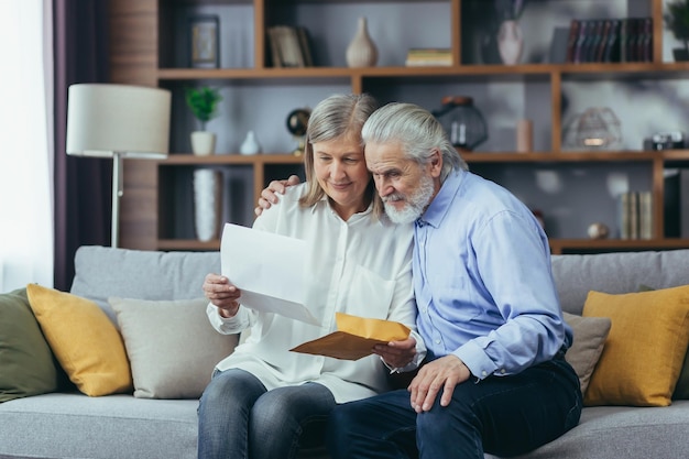 Happy senior retired couple sitting on sofa at home reading letter happy and smiling