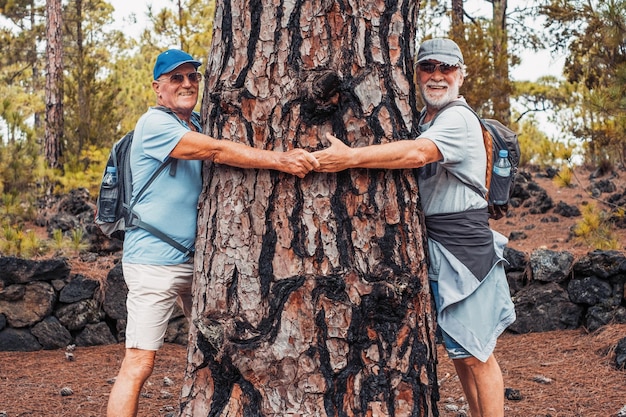 Happy senior retirees in mountain looking at camera laughing while embracing a large tree trunk