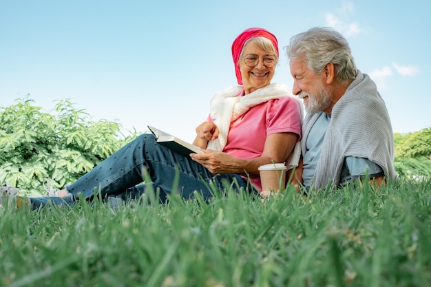 Happy senior smiling couple lying on the grass in public park with a coffee cup and a book