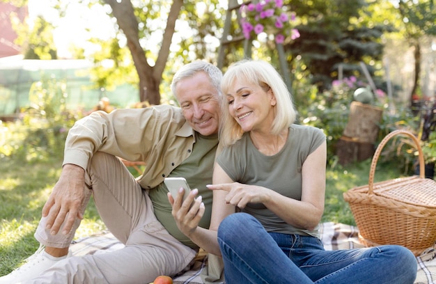 Happy senior spouses sitting on blanket during picnic in garden and using smartphone resting