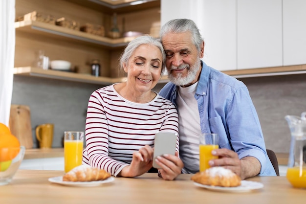 Happy Senior Spouses Spending Time With Smartphone While Having Breakfast In Kitchen