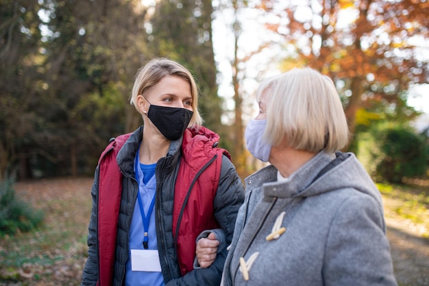 Happy senior woman and caregiver outdoors on a walk in park, coronavirus concept.