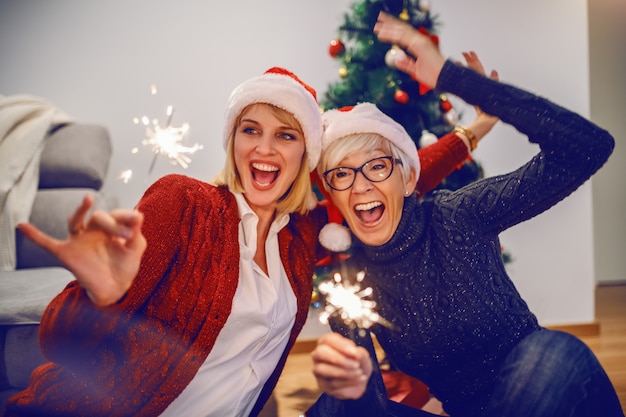 Happy senior woman and her daughter celebrating new year. Both having santa hats on heads and holding sparklers. In background is christmas tree. Family values concept.