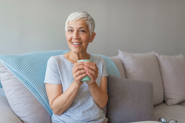 Happy senior woman sitting on a couch while she is drinking a cup of coffee.