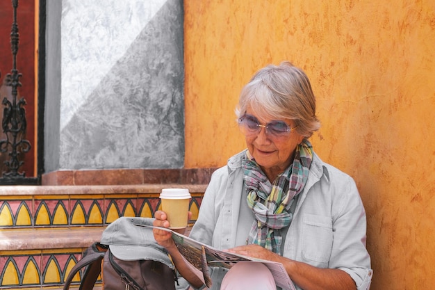 Happy senior woman sitting on a staircase consulting a map holding a takeaway coffee elderly traveler lady enjoying freetime or retirement