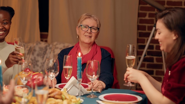 Happy senior woman sitting at table with relatives while enjoying Christmas dinner smiling at camera. Joyful elder person celebrating winter feast with close family members.