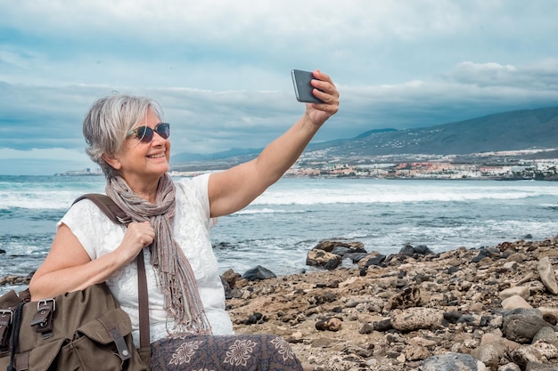 Happy senior woman with sunglasses enjoying travel and freedom at the beach using smartphone