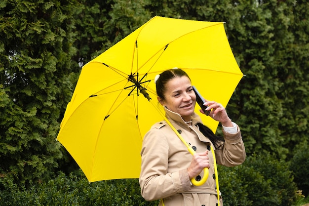 Happy senior woman in yellow rain coat with yellow umbrella is talking on a smartphone and walking