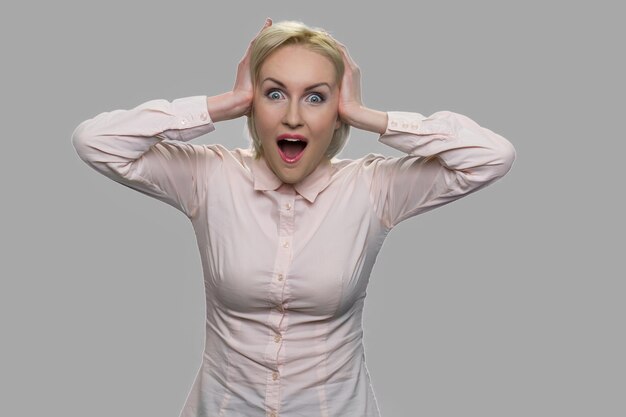 Happy shocked business woman holding hands on head. Young emotional business lady looking excited against gray background.