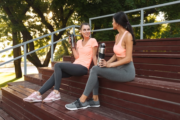 happy slim women in sportswear drinking water from bottles while sitting on bench in sports ground