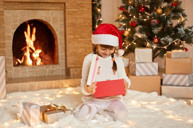 Happy small girl opening Christmas present box, looks inside with smile, being happy to get such gift, wearing white pullover and santa claus hat, sitting on floor in festive room with xmas decoration