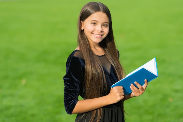 Happy small kid in school uniform smile holding study book green grass sunny summer outdoors education