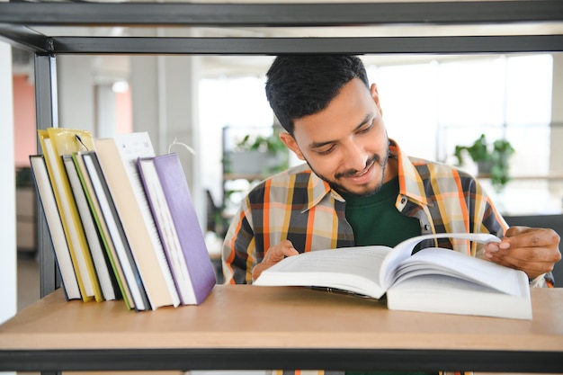 Happy smart indian or arabian guy mixed race male university student stands in the library against the background of bookshelves holds a lot of books in his hands looks at camera smiles friendly