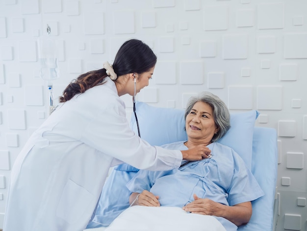 Happy smile Asian elderly old female patient in light blue dress lying on bed while beautiful young woman doctor in white suit using stethoscope to examining listen to her heartbeat in hospital room