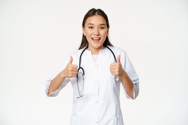 Happy smiling asian woman doctor showing thumbs up in approval satisfied with patient condition appr...