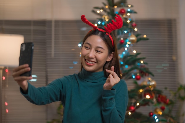 Happy smiling Asian woman video call via smartphone to celebrate with her friends or family during Christmas at home doing mini heart gesture