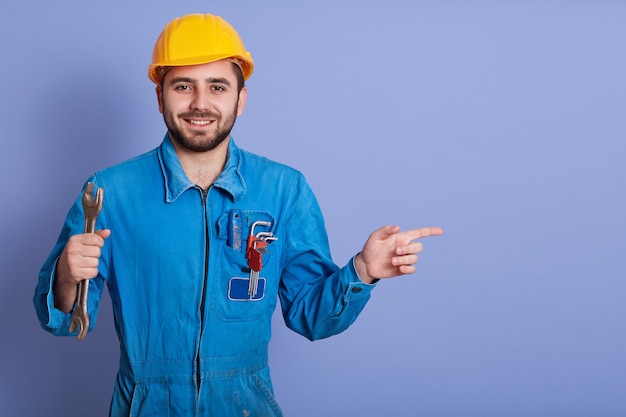 Happy and smiling bearded engineer with wrench tool in hand and pointing aside with index finger while stands against