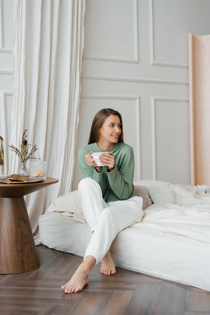 Happy smiling beautiful European woman enjoys the morning and inhales the aroma of a cup of coffee in her bedroom Enjoying free and relaxing time the concept of a lazy weekend