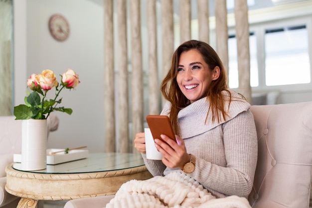 Happy smiling beautiful girl using smartphone device at cozy home , attractive young woman chatting with friends at social network while sitting on sofa at home and drinking coffee