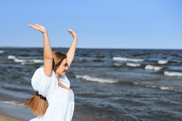 Happy smiling beautiful woman on the ocean beach standing in a white summer dress, raising hands