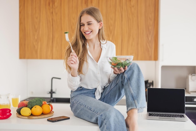 Happy and smiling blonde woman sitting on top of kitchen table and eating homemade salad laptop with copy space