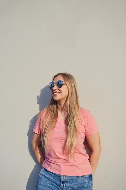 Happy smiling blonde woman in sunglasses in a pink tshirt standing against a gray wall on a sunny day