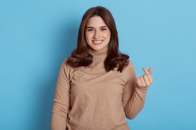 Happy smiling Caucasian woman makes korean hand sign, expresses love, makes like gesture, dressed in casual beige turtleneck, isolated on beige wall.