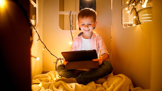 Happy smiling cheerful boy watching cartoons and playing games on tablet computer at night. Concept of child education and studying at night.