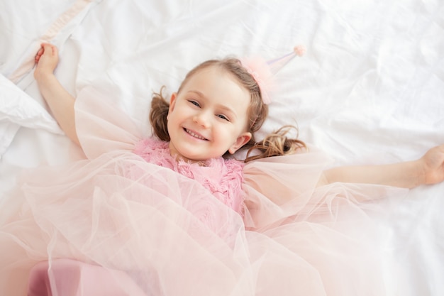 happy smiling child girl in pink dress and birthday cap lies on bed spread arms photo above