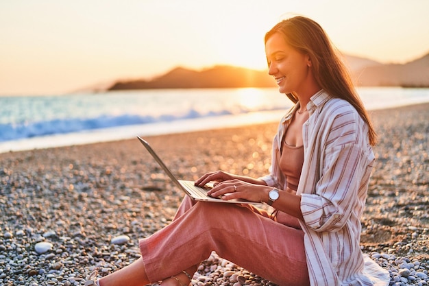 Happy smiling cute joyful satisfied girl freelancer remote working online on the seashore at sunset time Work everywhere