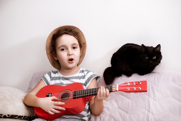 Happy smiling cute little girl in hat singing and playing small guitar ukulele at home Funny black cat at background listening