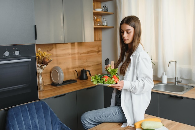 Happy smiling cute woman is preparing a fresh healthy vegan salad with many vegetables in the kitchen at home and trying a new recipe