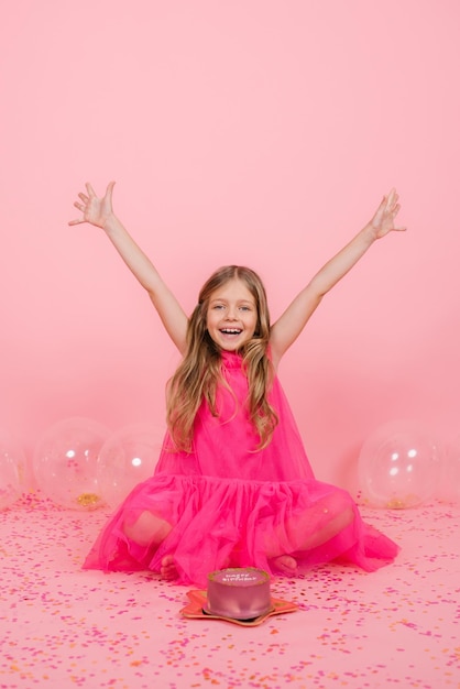 Happy smiling eight year old Caucasian girl made a wish near a bento cake for her birthday joyfully raised her hands to the sides sitting near balloons on a pink background with confetti