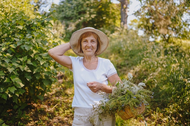 Happy smiling elderly senior woman in straw hat having fun posing in summer garden with flowers in