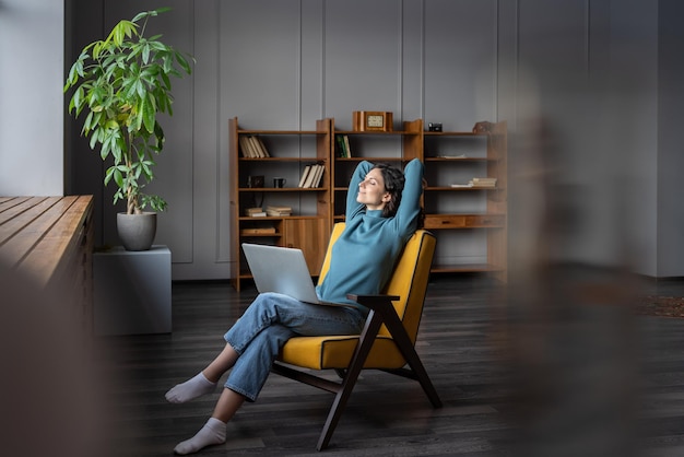 Happy smiling female freelancer with laptop on knees relaxing at home office with hands behind head