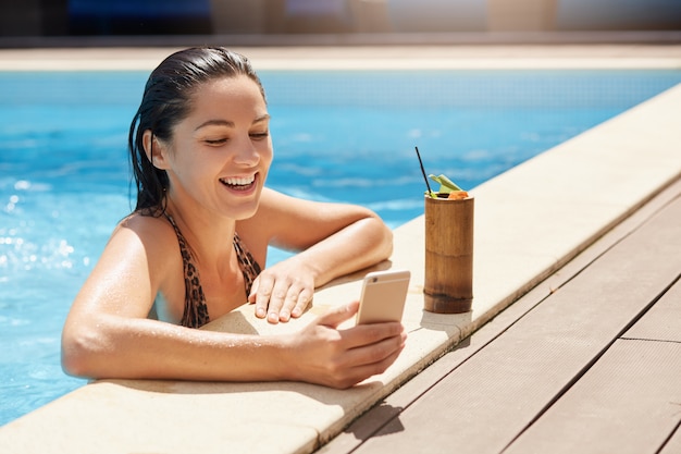 Happy smiling female with wet hair holding modern smart phone in hand, relaxing at poolside and drinking non alcoholic cocktail