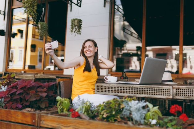 Happy smiling girl in outdoors street coffee shop cafe sitting at table with laptop pc computer doing taking selfie shot on mobile phone in restaurant during free time