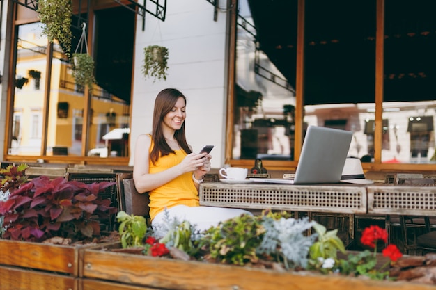 Happy smiling girl in outdoors street coffee shop cafe sitting at table with laptop pc computer, texting message on mobile phone friend, in restaurant during free time